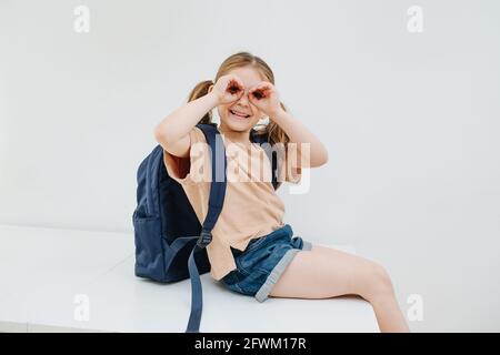 Dummes kleines Mädchen mit Spaß, Grimassen, googles Geste. Sie sitzt auf einem Tisch und schaut auf die Kamera. Auf weißem Hintergrund. Stockfoto