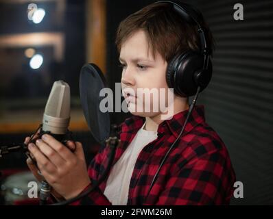 Junge im Karomudio und Kopfhörer singt oder spricht im Studio mit POP-Filter ins Mikrofon. Stockfoto