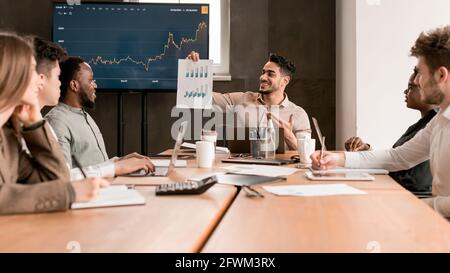 Mitarbeiter zeigt Papier mit Diagramm und sitzt am Schreibtisch Stockfoto