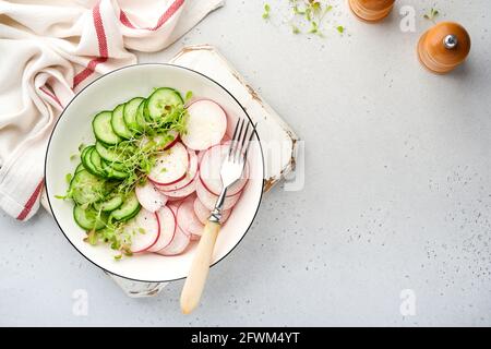 Frischer Salat mit rotem Rettich, Gurke, Gemüse, mikrogrünen Radieschen auf weißem Teller auf grauem Steingrund. Blick von oben. Konzept vegan und hea Stockfoto