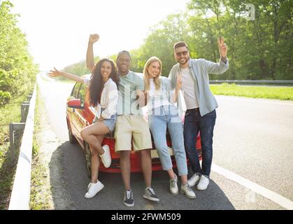 Porträt glücklicher, vielfältiger Freunde, die in der Nähe ihres Autos auf der Autobahn posieren, verschiedene Gesten machen und die Kamera anlächeln Stockfoto