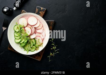 Frischer Salat mit rotem Rettich, Gurke, Gemüse, mikrogrünen Radieschen auf weißem Teller auf grauem Steingrund. Blick von oben. Konzept vegan und hea Stockfoto