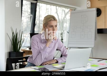 Durchdachte Geschäftsfrau, die mit einem Laptop am Tisch sitzt und über die Strategie nachdenkt Stockfoto