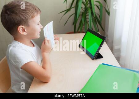 Lächelnder Schuljunge sitzt am Schreibtisch und schaut auf das Tablet mit grünem Bildschirm. Glückliches Kind zeigt sein Bild. Online-Unterricht aus der Ferne. Grüne Chroma-Taste auf dem Monitor. Seitenansicht Stockfoto