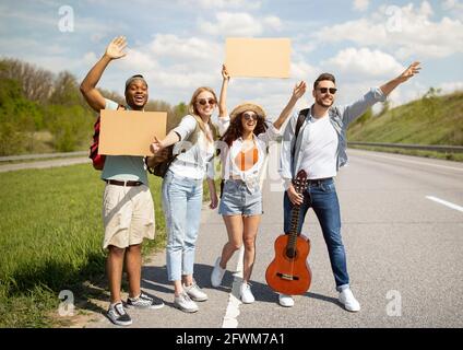 Glückliche, vielfältige Freunde stehen auf der Autobahn mit leerem Schild, winken mit dem Auto, gehen gemeinsam auf Reise, im Freien Stockfoto