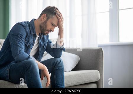 Nachdenklicher Mann, der zu Hause auf der Couch sitzt, Seitenansicht Stockfoto