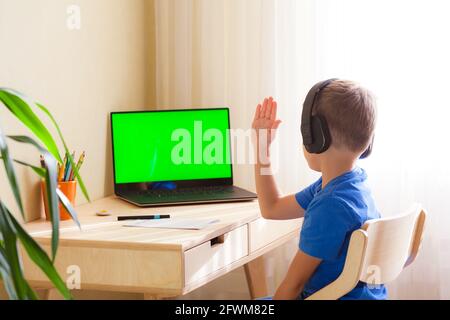 Ein intelligenter Schuljunge mit Kopfhörern sitzt am Tisch und schaut auf den Computer. Ein Kind zieht an der Hand, um zu antworten. Online-Unterricht aus der Ferne. Grüne Chroma-Taste auf dem Monitor. Stockfoto