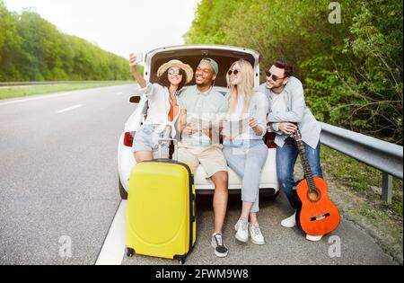 Eine Gruppe von besten Freunden, die während der Autoreise die Karte überprüfen, im Kofferraum sitzen und Selfie auf ihrer Reise machen. Anhalter Stockfoto