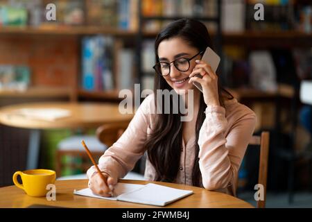 Glückliche arabische Dame, die auf dem Smartphone spricht und sich in der Cafeteria Notizen macht, ihren Zeitplan verwaltet oder Meetings plant. Stockfoto