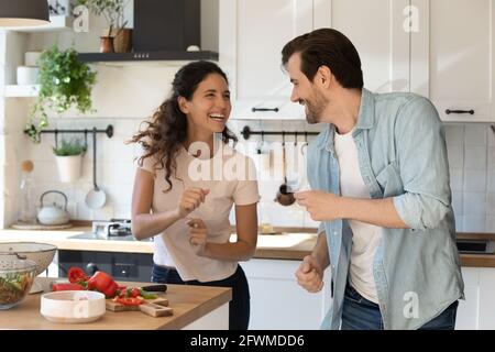 Glückliches Millennial-Familienpaar, das zu energiegeladener Musik in der Küche tanzt. Stockfoto