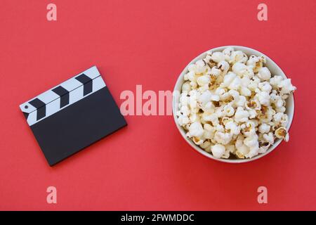 Schüssel Popcorn und Filmklapper isoliert auf rotem Hintergrund. Draufsicht. Fettige und ungesunde Snacks. Filmzeit. Unterhaltungsbranche. Minimalistischer Rücken Stockfoto
