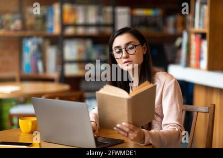 Nachdenkliche lateinerin liest vor dem Laptop in der Cafeteria ein Buch, studiert und bereitet sich auf die Prüfung vor. Stockfoto