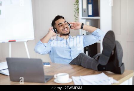 Pause von der Arbeit. Lächelnder arabischer Mann, der mit Kopfhörern Musik hört und sich am Arbeitsplatz entspannt Stockfoto