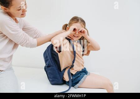 Junge Mutter, die ihrer Tochter hilft, sich für die Schule vorzubereiten, ist das Mädchen gelangweilt, hat dummen Spaß, Grimassen, macht googles Geste. Auf weißem Hintergrund. Stockfoto