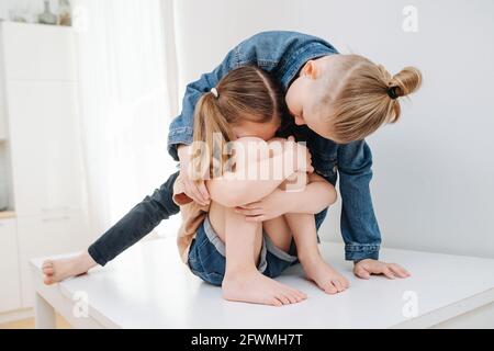 Weinendes kleines Mädchen, das auf einem Tisch sitzt. Ihr Bruder versuchte, sie zu beruhigen, um Frieden zu schließen. Sie hat ihren Rücken gekehrt, ihre Knie umarmt und geschmürt. Stockfoto