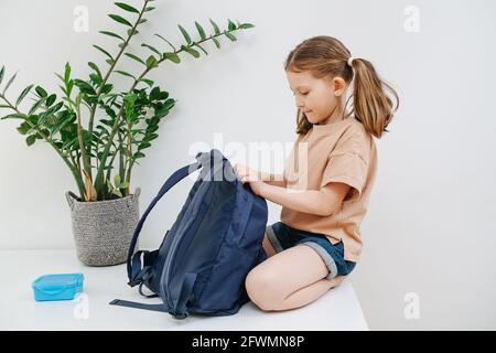 Das verantwortliche Schulmädchen sitzt auf einem Tisch und zippt ihren Rucksack. Stockfoto