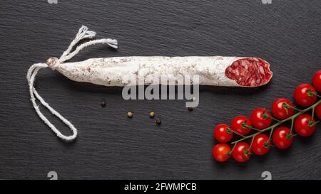 Dünne katalanische Fuetwurst und ein paar rote Kirschtomaten auf einer schwarzen Schiefer-Oberfläche. Breites Bild der traditionellen spanischen trockengeräucherten Schweinefleischsalami. Stockfoto