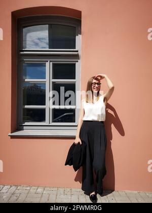 Eine junge Geschäftsfrau in voller Höhe hält eine Jacke in der Nähe Die orangefarbene Wand und das Fenster Stockfoto