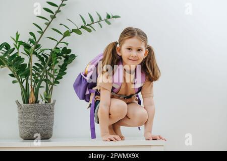 Verspieltes kleines Schulmädchen auf einem weißen Tisch mit purpurem Rucksack. Überweiß Stockfoto