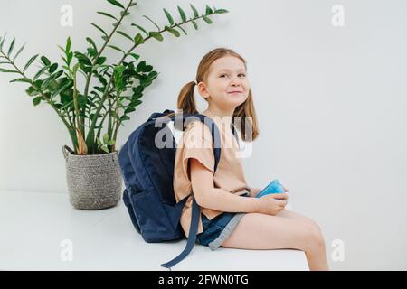 Kleines Schulmädchen, das auf einem Tisch sitzt und jemanden anlächelt. Weißer Hintergrund Stockfoto