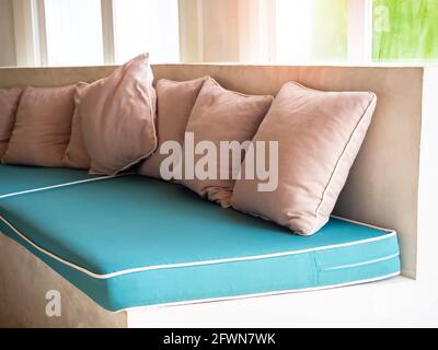 Viele beigefarbene Kissen auf grünen Sitzkissen auf einem Betonsofa in der Nähe des Glasfensters. Stockfoto
