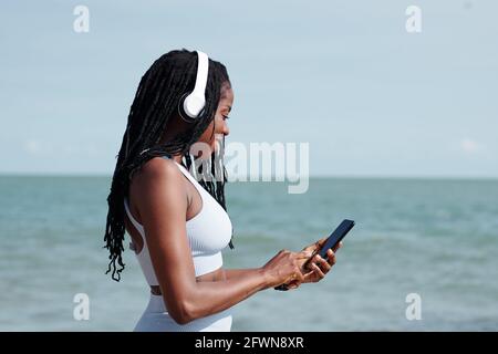 Lächelnd, fit, junge Frau mit Kopfhörern, die Musik für sie auswählt Traning über Anwendung auf dem Smartphone Stockfoto