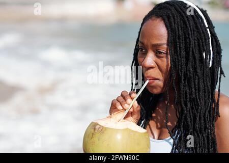 Porträt einer lächelnden jungen Frau mit Dreadlocks, die Musik hört Mit Kopfhörern und Kokosnusscocktail Stockfoto