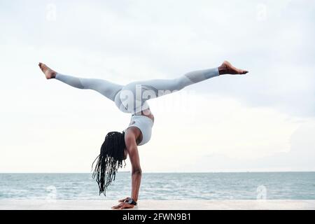 Seitenansicht einer athletischen jungen Frau, die Yoga praktiziert und am Strand einen Handstand mit Splits Pose macht Stockfoto