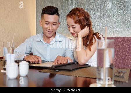 Lächelnder junger Freund und Freundin sitzen am Restauranttisch und Lesen Sie die Speisekarte, bevor Sie Lebensmittel bestellen Stockfoto