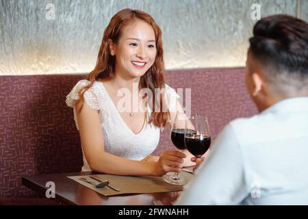 Schöne lächelnde junge Frau, die mit ihrem Freund am Restauranttisch sitzt Und ein Glas Rotwein trinken Stockfoto