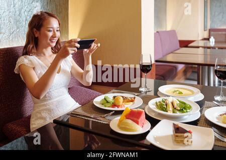 Ziemlich lächelnde junge Frau fotografiert Restauranttisch mit verschiedenen Gerichten Für soziale Medien Stockfoto