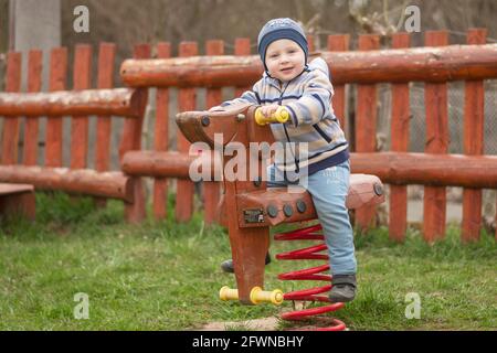 Der zweijährige kaukasische Junge schaukelt auf einer Schaukel auf dem Spielplatz. Zeigen Sie die Kamera an. Stockfoto