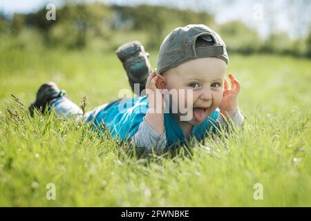 Lustiges Gesicht. Der zwei Jahre alte kaukasische Junge mit Baseballmütze liegt im Gras mit Zungenzunge. Stockfoto
