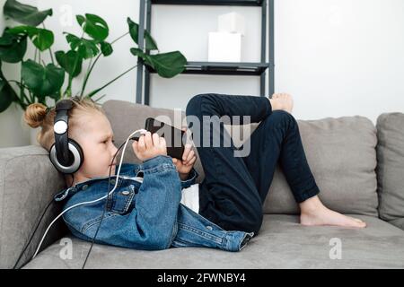 Fokussierter Junge mit Kopfhörern, der sein Telefon auf einer Couch beobachtete. Seitenansicht. Stockfoto