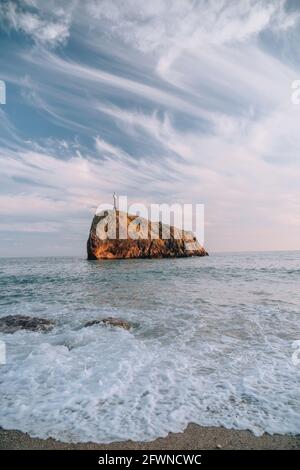 Heller Sonnenuntergang am Meer. Die Wellen stürzen in den Felsen, erleuchtet von dem warmen Sonnenuntergang, Sand und Kieselsteinen, vulkanischem Basalt wie in Island. Meereswelle bricht in Stockfoto