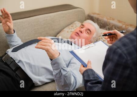 Ein älterer Mann liegt während eines Termins mit einem Psychotherapeuten auf einer Couch. Ein älterer männlicher Patient in einer individuellen Konsultation mit einer Frau Stockfoto