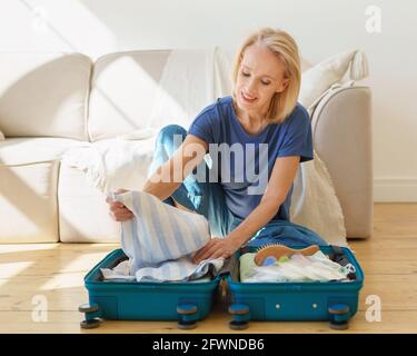 Nachdenkliche, reife Frau aus den 60ern, die Koffer packte und das Gepäck für eine Reise vorbereitete, während sie zu Hause auf dem Holzboden saß. Ältere Dame, die sich auf den Sommerurlaub vorbereitet. Das Konzept des Reisens im Ruhestand Stockfoto