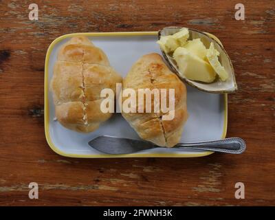 Zwei frische Croissants auf der Schüssel mit Butter in Tasse und Messer, französisches Brot auf braunem Holztisch Stockfoto