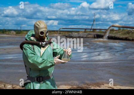 Ein männlicher Umweltschützer in einem grünen Schutzanzug und einer Gasmaske nimmt eine Wasserprobe in einem verschmutzten See für die Forschung. Abfallproduktion. Wissenschaftler Stockfoto