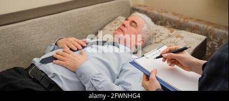 Ein älterer Mann liegt während eines Termins mit einem Psychotherapeuten auf einer Couch. Ein älterer männlicher Patient in einer individuellen Konsultation mit einer Frau Stockfoto