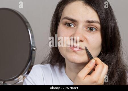 Eine Frau vor einem Spiegel, die Haare über ihrer Oberlippe zupfen. Den Schnurrbart mit der Pinzette selbst entfernen Stockfoto