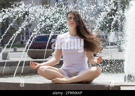 Schöne Brünette Frau in weißen Kleidern meditiert in der Nähe eines Brunnens auf der Straße in der Stadt. Yoga und Meditation, verlangsamt sich im Rhythmus des Modus Stockfoto