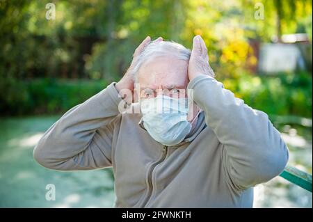 Kranker, trauriger grauhaariger alter Mann sitzt in einer medizinischen Maske auf der Straße. Der Mann hat starke Kopfschmerzen. Das Konzept der Kopfschmerzen. Frühjahrsallergie, Methoden der Stockfoto