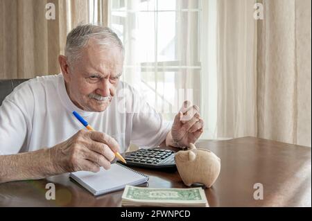 Der alte Mann mit Bleistift zählt Pfennige aus der Pensionskasse, die an einem Tisch im Zimmer sitzt. Besorgter älterer Mann steckt Geld in eine Sparschwein. Rentner c Stockfoto