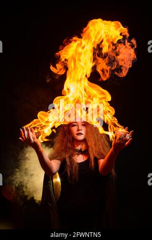 Eine Hexe mit langen lockigen Haaren hält ein magisches Feuer. Flammenzungen auf den Handflächen einer rothaarigen Frau. Stockfoto