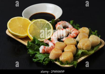 Gebratener Jakobsmuschel mit Garnelenschwänzen mit Zitrone auf Salatblättern auf einem Holzteller mit Sauce. Selektiver Fokus. Stockfoto