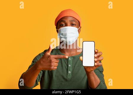 Junger schwarzer Mann in medizinischer Maske zeigt auf Smartphone mit Weißer Bildschirm Stockfoto