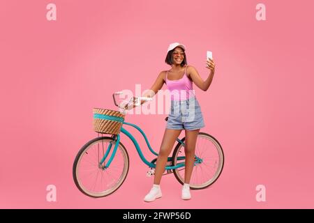 Volle Länge der glücklichen schwarzen Dame in Sommerkleidung nehmen Selfie auf dem Smartphone neben dem klassischen Fahrrad auf rosa Hintergrund Stockfoto