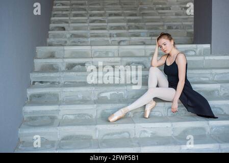 Ballerina in tutu posiert sitzen auf der Treppe EINE schöne Junge Frau in einem schwarzen Kleid und spitzenschuhe ist In einer eleganten Pose auf dem sitzen Stockfoto