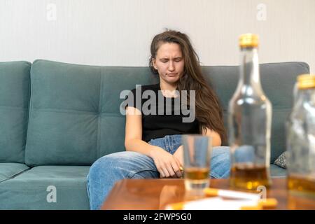 Die junge dunkelhaarige Frau sitzt mit geschlossenen Augen auf dem Sofa, neben leeren Flaschen, Zigaretten. Gesellschaftliches Problem des Alkoholismus Stockfoto